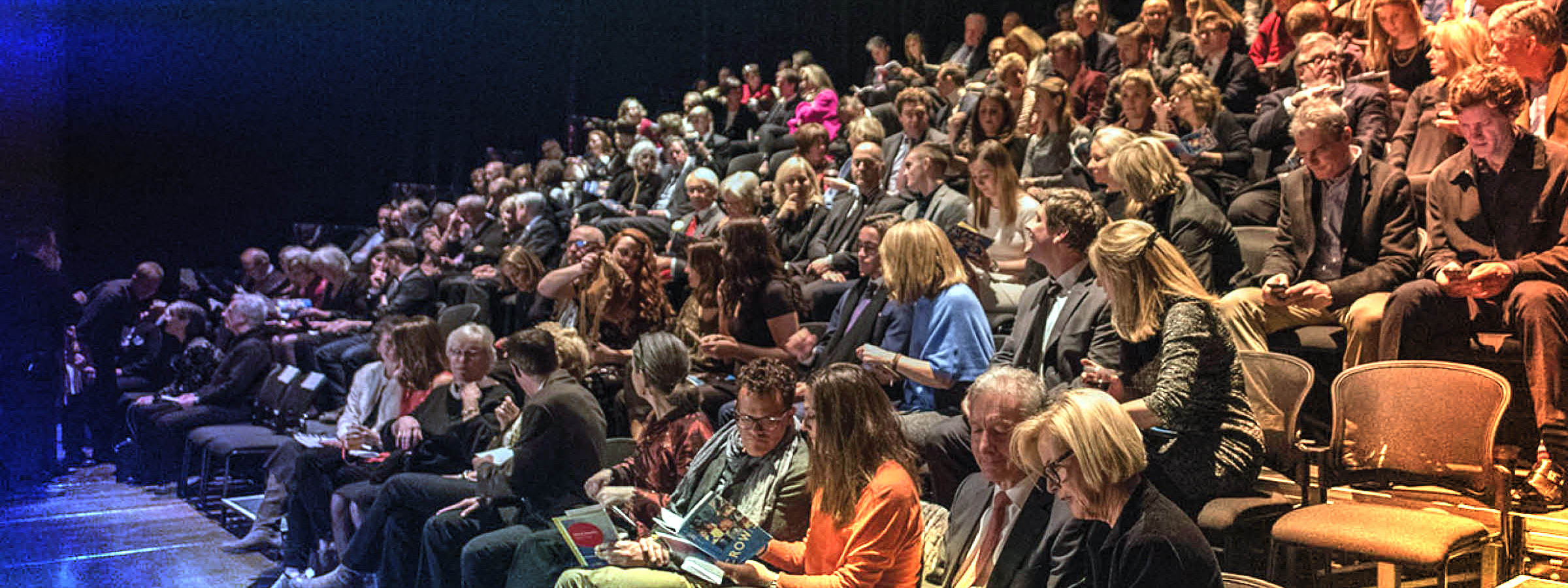 A large group of people seated in tiered rows inside a theatre. Most individuals are dressed in formal or semi-formal attire and are engaged in various pre-show activities such as reading programs, chatting, or using their phones. The seating area is nearly full, with only a few empty chairs visible near the front.