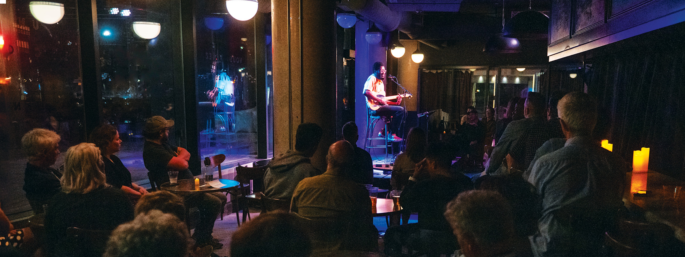 A person is performing on a small stage in a dimly lit venue, seated on a tall stool and playing an electric guitar while singing into a microphone. The performer is illuminated by stage lighting, while a seated audience watches attentively from small tables arranged throughout the space. Large windows line the left side of the room, reflecting the interior lights and showing faint views of the outside. The atmosphere is intimate and relaxed, with warm lighting and candles on some tables.