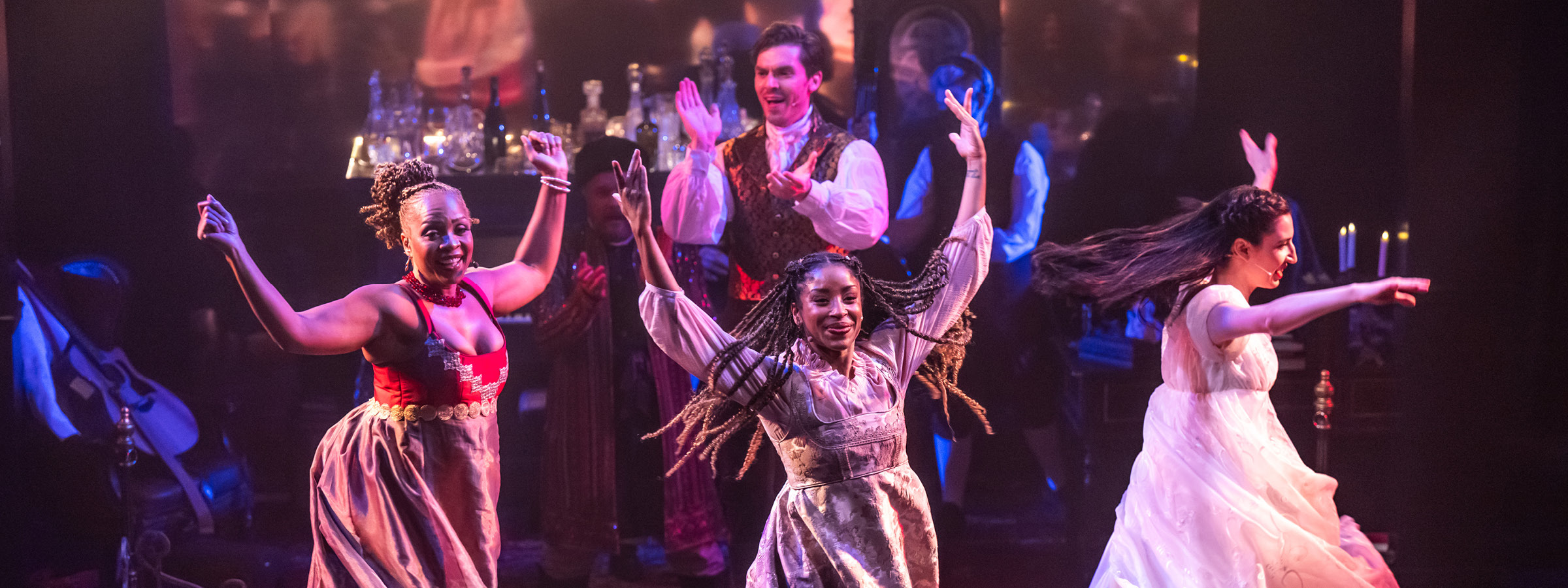 Three women in period costumes are dancing with raised arms and joyful expressions on a dimly lit stage. The woman on the left wears a red and gold bodice with a flowing beige skirt, the centre dancer has long braids and a silver brocade dress over a blouse, and the dancer on the right wears a flowing white dress. A man in a white shirt and ornate vest claps enthusiastically in the background, surrounded by others in costume and a backdrop featuring candles and bottles.