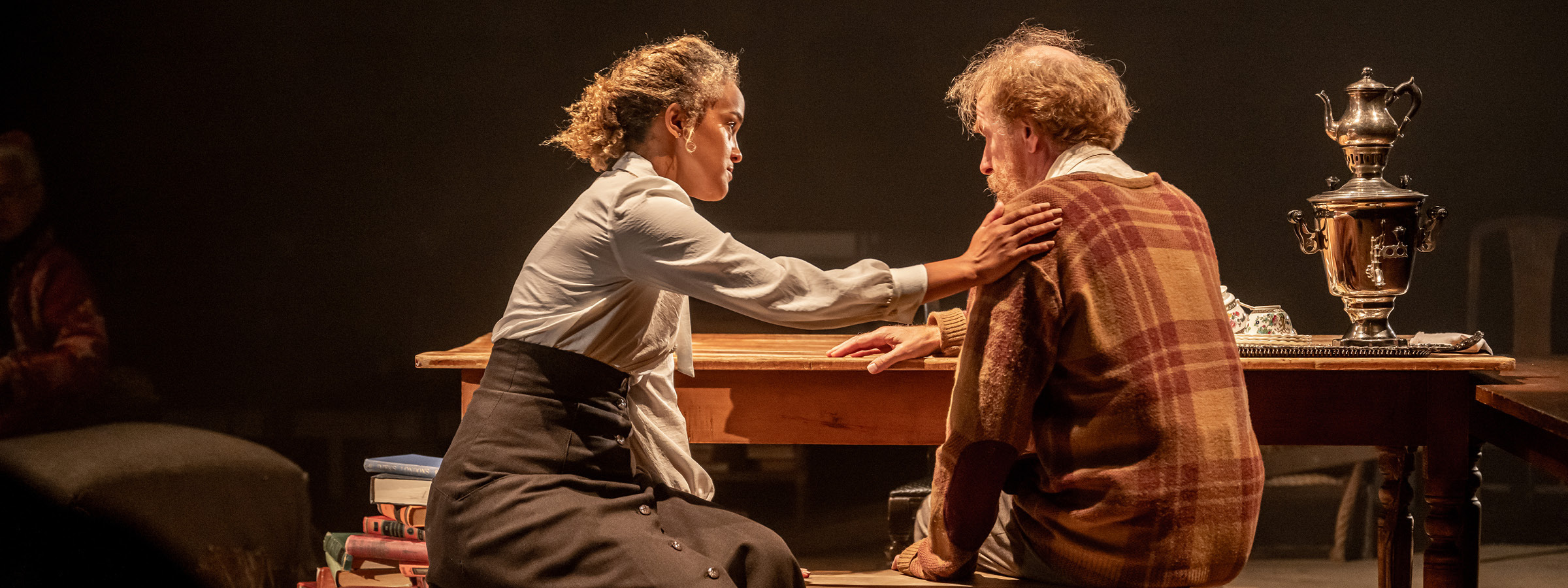 A theatrical scene with two actors at a wooden table under warm stage lighting. A woman in a white blouse and dark skirt sits on the left, leaning forward with a hand placed gently on the shoulder of an older man in a plaid sweater seated to the right. The man looks downcast, and the woman appears comforting. On the table are stacked books, a decorative tea set, and an ornate metal samovar. The background is dimly lit, with another person partially visible on the left.