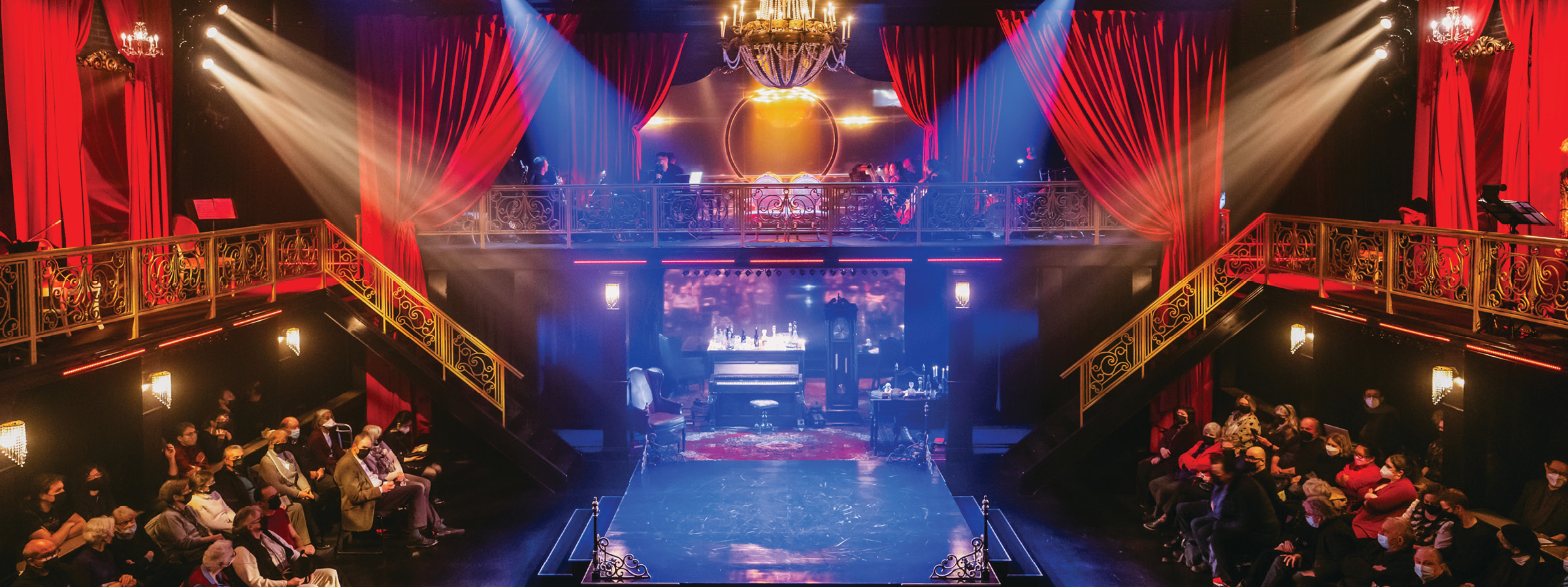 Interior of a theatre with ornate gold railings, red velvet drapes, and a grand chandelier hanging above a central stage. The stage is set with vintage furniture including a piano, chairs, and a grandfather clock. An audience is seated on both sides in front of the stage, with some people wearing face masks. Beams of stage light illuminate the scene from above, and additional audience members can be seen on a balcony level in the background.
