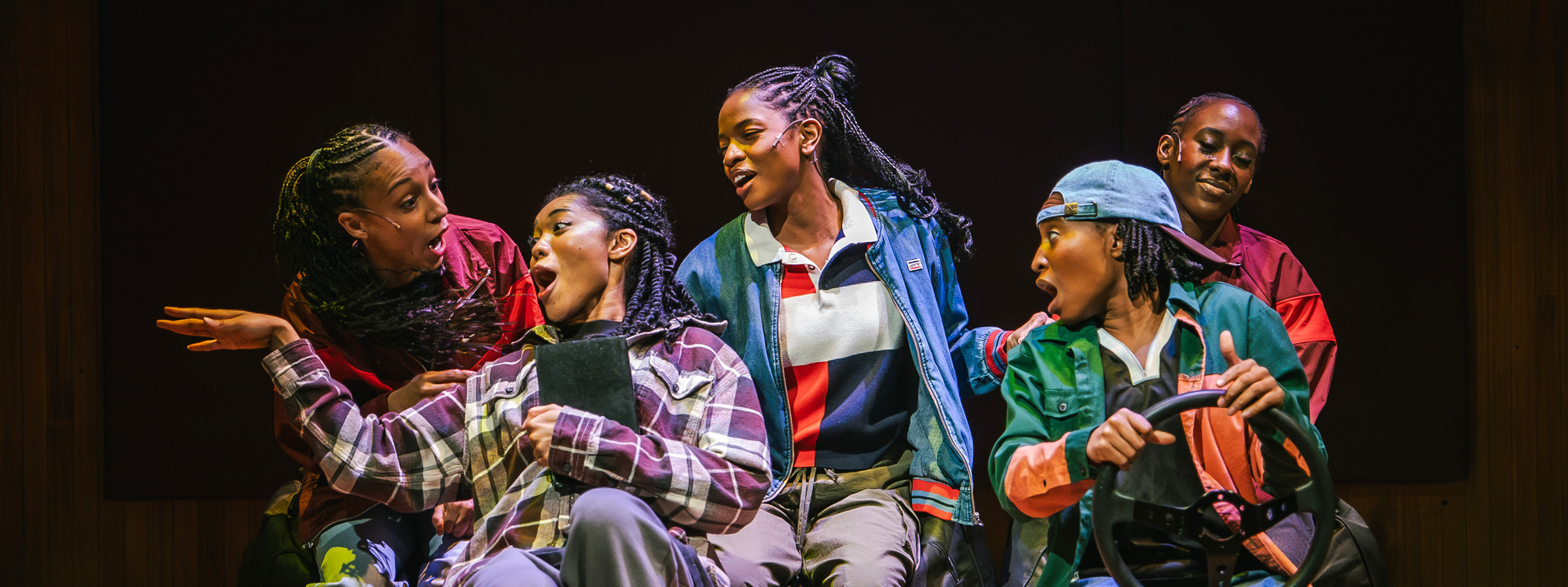 Five performers are on stage, animatedly interacting with each other. The central figure in a plaid shirt has an exaggerated surprised expression, while the person to their right holds a steering wheel, suggesting a driving scene. The others are dressed casually in layered outfits and appear engaged in lively conversation or storytelling, with expressive faces and gestures.
