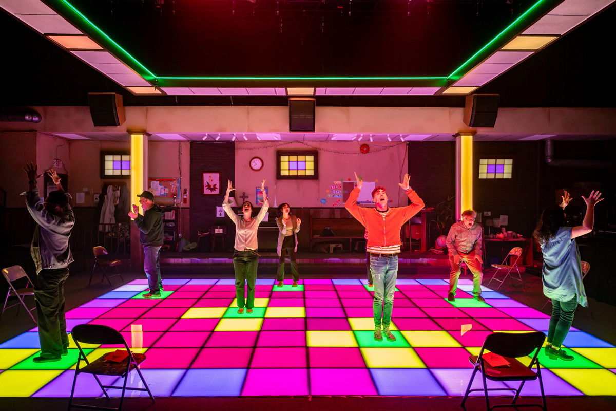 A colourful stage with a glowing neon grid floor in pink, yellow, and purple squares. Seven performers are spread out across the stage, raising their arms in expressive gestures. The setting resembles a community centre or meeting space, with folding chairs and wall decorations in the background.