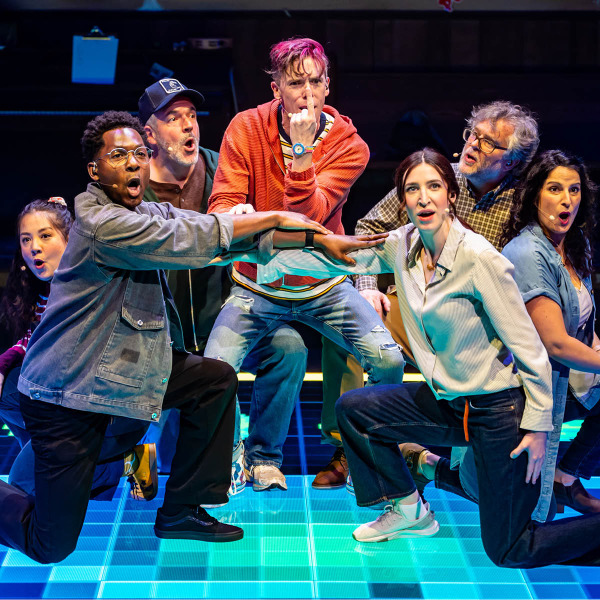Seven actors are performing on a brightly lit, glowing blue grid stage. They are in energetic, expressive poses, with some kneeling and pointing forward. The actor at the centre has pink hair, wears a red hoodie and ripped jeans, and is holding his index finger vertically in front of his nose while making a serious facial expression. The others show animated expressions, suggesting a moment of intensity or high emotion in the performance.aptured mid-song or mid-expression, against a dark background with blue lighting. The central figure, a woman with long dark hair, appears joyful with her eyes closed and head slightly tilted back. The surrounding individuals show a range of emotions, including serenity, intensity, and passion, suggesting a musical or choral performance.