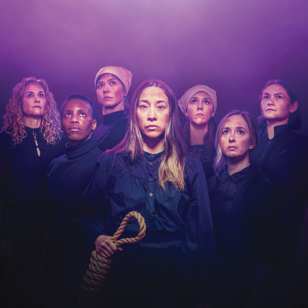 A dramatic portrait of seven women dressed in dark, somber clothing, standing closely together under purple lighting. The central woman holds a noose in her hand and gazes directly at the camera with a solemn expression. The others look upward or into the distance, conveying a sense of defiance, tension, or resolve.