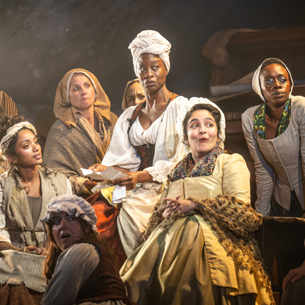 Women dressed in historical 18th-century-style clothing are gathered closely together in what appears to be a dimly lit theatrical setting. Most of them wear bonnets, shawls, and layered dresses in muted earth tones. The woman in the centre, wearing a white headwrap and holding a piece of paper, looks directly ahead with a serious expression. The others around her show a mix of curiosity, surprise, and attentiveness, suggesting an intense or dramatic moment in the performance.