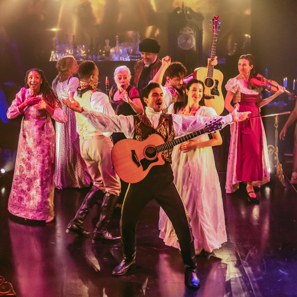 A vibrant stage scene featuring a group of performers in early 19th century Russian garb, energetically singing and playing instruments. The central figure, a man in a brocade vest, holds a guitar and sings passionately with arms outstretched. Other performers dance, sing, and play instruments including a violin and another guitar. The background is warmly lit with candles and antique props, evoking a festive, theatrical atmosphere.