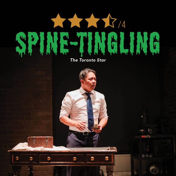 A man in a white dress shirt and blue tie stands behind a wooden desk on a dimly lit stage, holding a small object in each hand. On the desk are a cloth-covered surface and a small wooden chest. Behind him is a dark backdrop, and to the side is industrial-looking equipment. Above the scene, there is a graphic with four and a half yellow stars and green text reading 