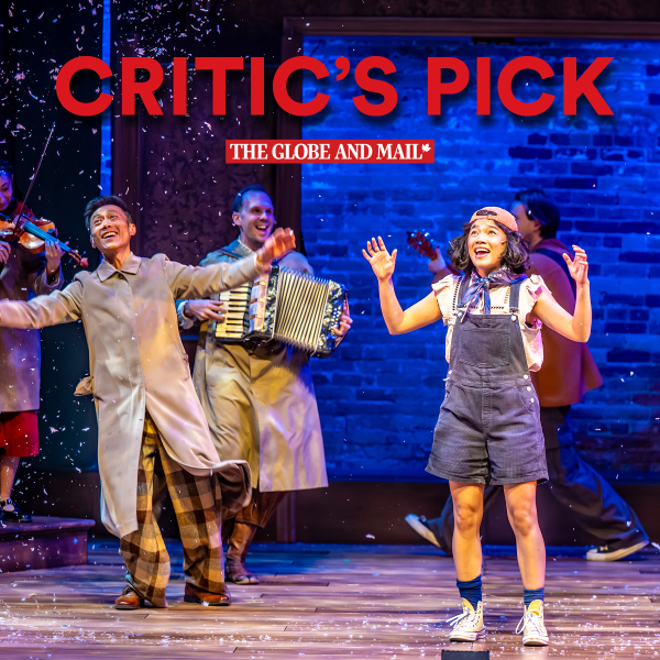 A smiling man in a long beige coat and plaid pants dances with arms outstretched, while another man behind him plays an accordion. To the right, a young woman in overalls, high socks, and a backward cap stands with hands raised and mouth open in excitement. Other performers in the background play instruments, including a violin and a guitar. Snow-like confetti falls around them. The backdrop is a brick wall lit with blue lighting. At the top, bold red text reads “CRITIC’S PICK” with “THE GLOBE AND MAIL” logo beneath it.