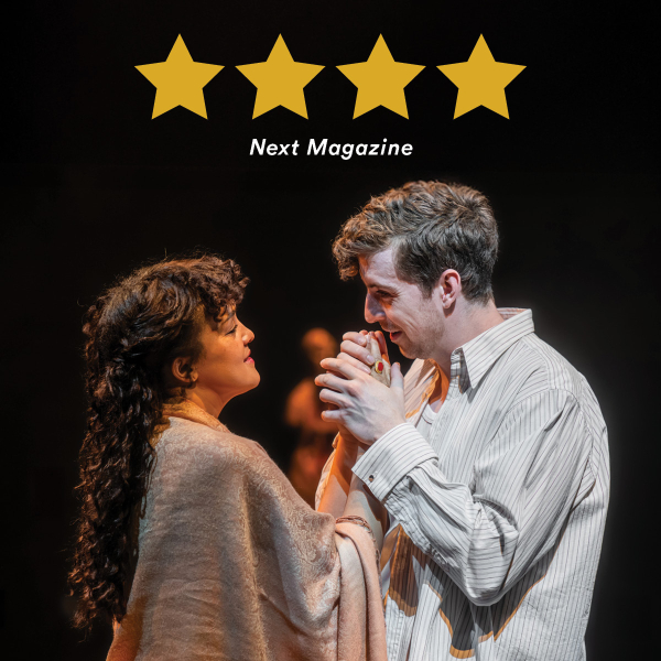Four gold stars across the top with the white text “Next Magazine” beneath them, above a photo of two people facing each other on a dimly lit stage. A person with long, dark curly hair and a light shawl smiles up at a man in a striped button-up shirt; he holds their hands near his mouth and looks emotional. Blurred figures and warm spotlights appear in the dark background.