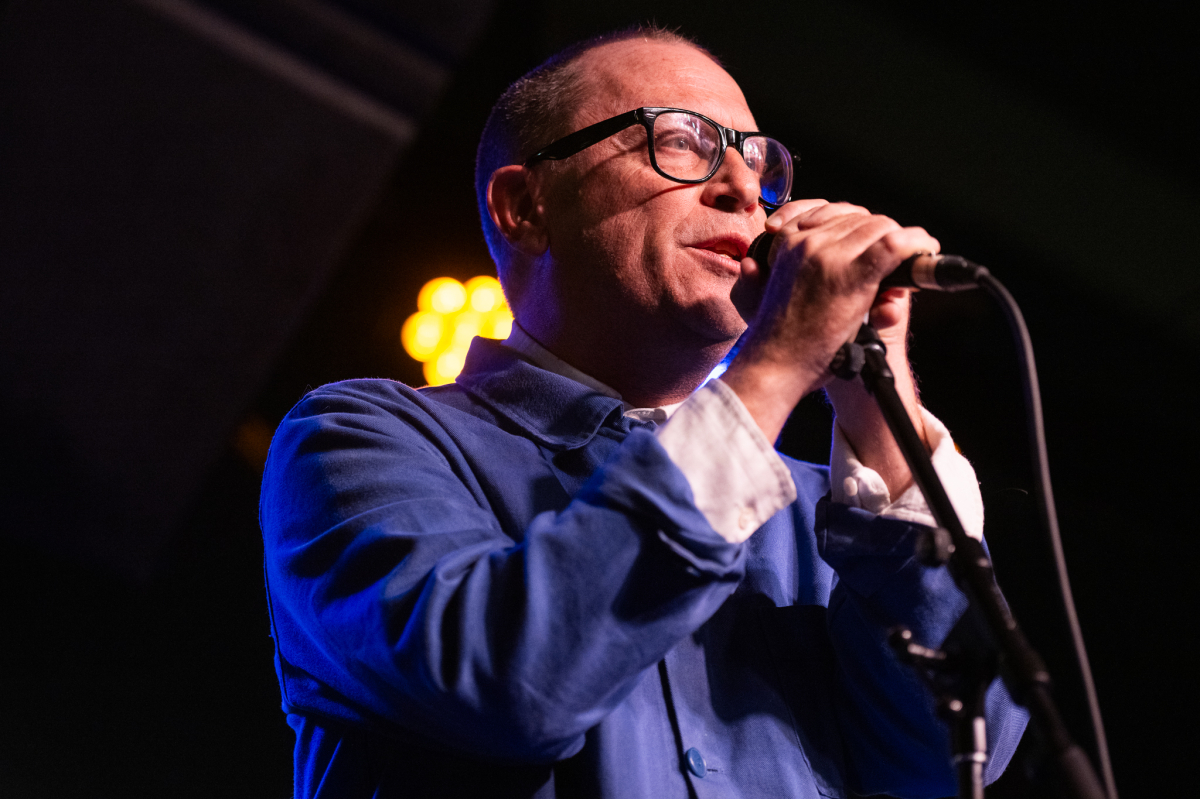 A man wearing glasses and a blue jacket is holding a microphone and speaking or singing into it on stage. He is lit by stage lighting with a visible warm yellow light behind him and blue-toned lighting on his clothing. A microphone stand is visible in the foreground.