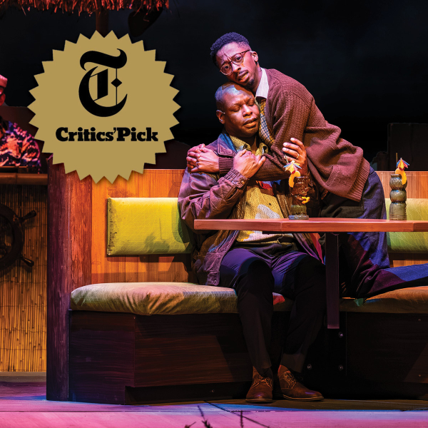 Two actors embrace in a theatre booth beside drinks with umbrella garnishes, with a gold New York Times Critics’ Pick seal on the left.