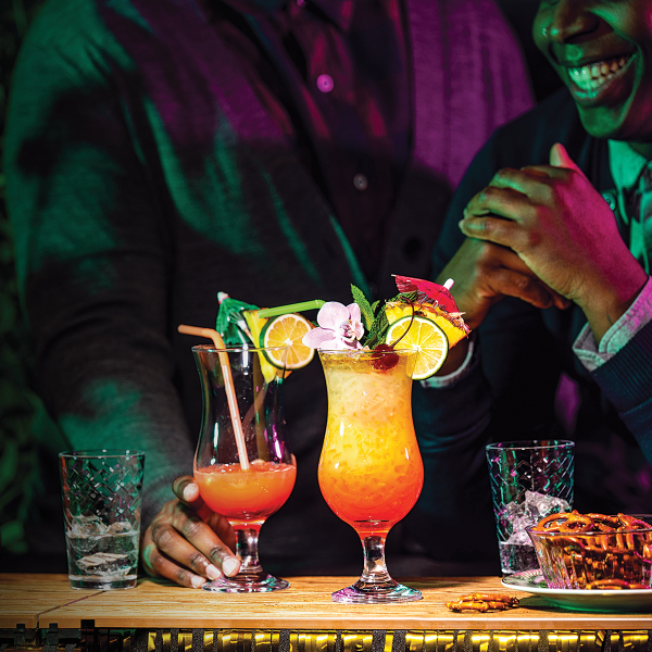 Two people at a bar or restaurant, partially visible and lit with colourful lighting, sit in front of a wooden surface with two elaborate cocktails garnished with citrus slices, cherries, mint, umbrellas, and a flower. There are also glasses of ice water and a bowl of pretzels on the table. One person is smiling broadly with clasped hands, while the other has a hand resting near a drink. The background includes plants and decorative lighting.