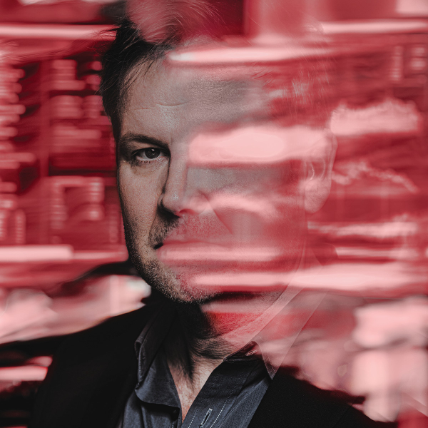 Close-up portrait of a dark-haired man with short facial hair wearing black, seen through layers of translucent red reflections and horizontal streaks that partially obscure his face. Blurred shelves filled with cylindrical objects appear in the background.