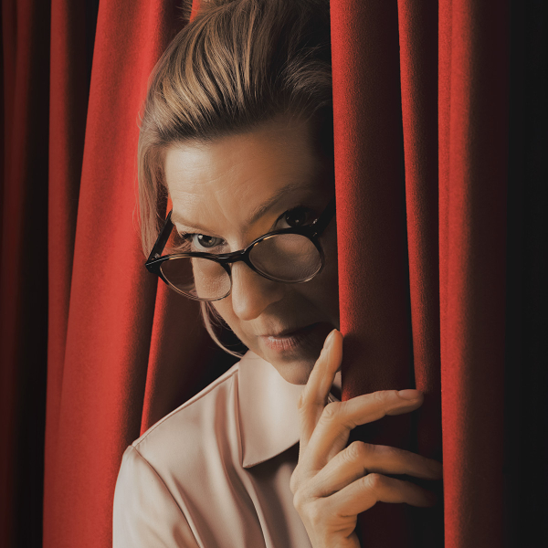 Close-up of a woman wearing dark-rimmed glasses and a pale collared blouse, peering through a narrow opening between deep red theatre curtains. One hand rests against the curtain as warm light falls across her face and hair.