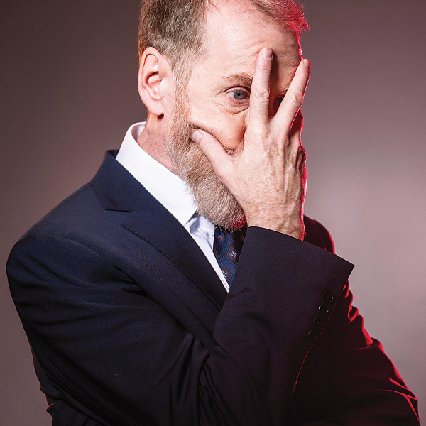 Close-up portrait of a bearded man in a dark suit, white shirt, and patterned tie, covering much of his face with one hand while looking through his fingers toward the camera. Red light highlights one side of his head and jacket against a plain grey background.