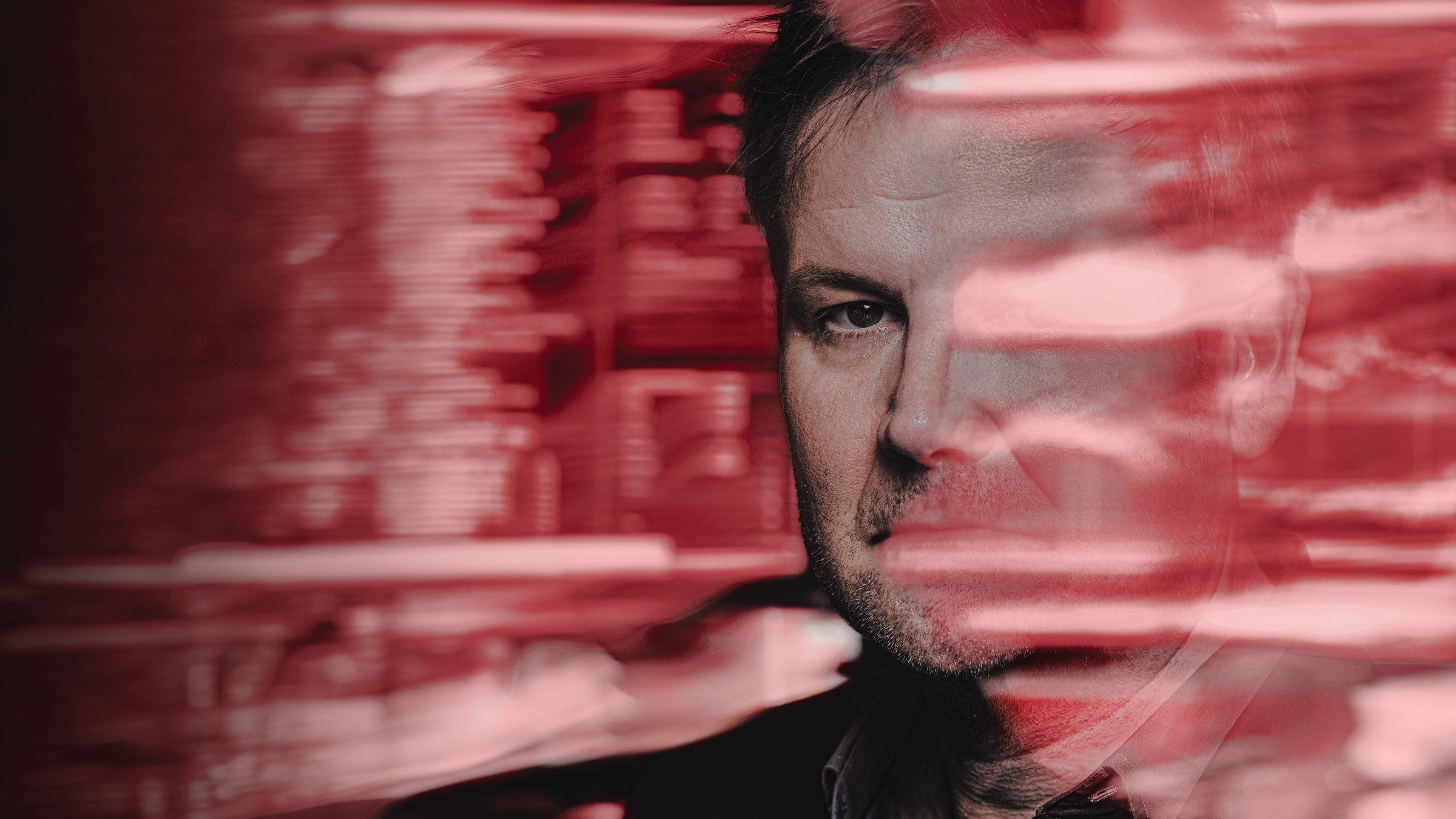 Close-up portrait of a man positioned on the right, looking directly at the camera, with blurred red and pale pink horizontal streaks layered across his face and the dark red background.