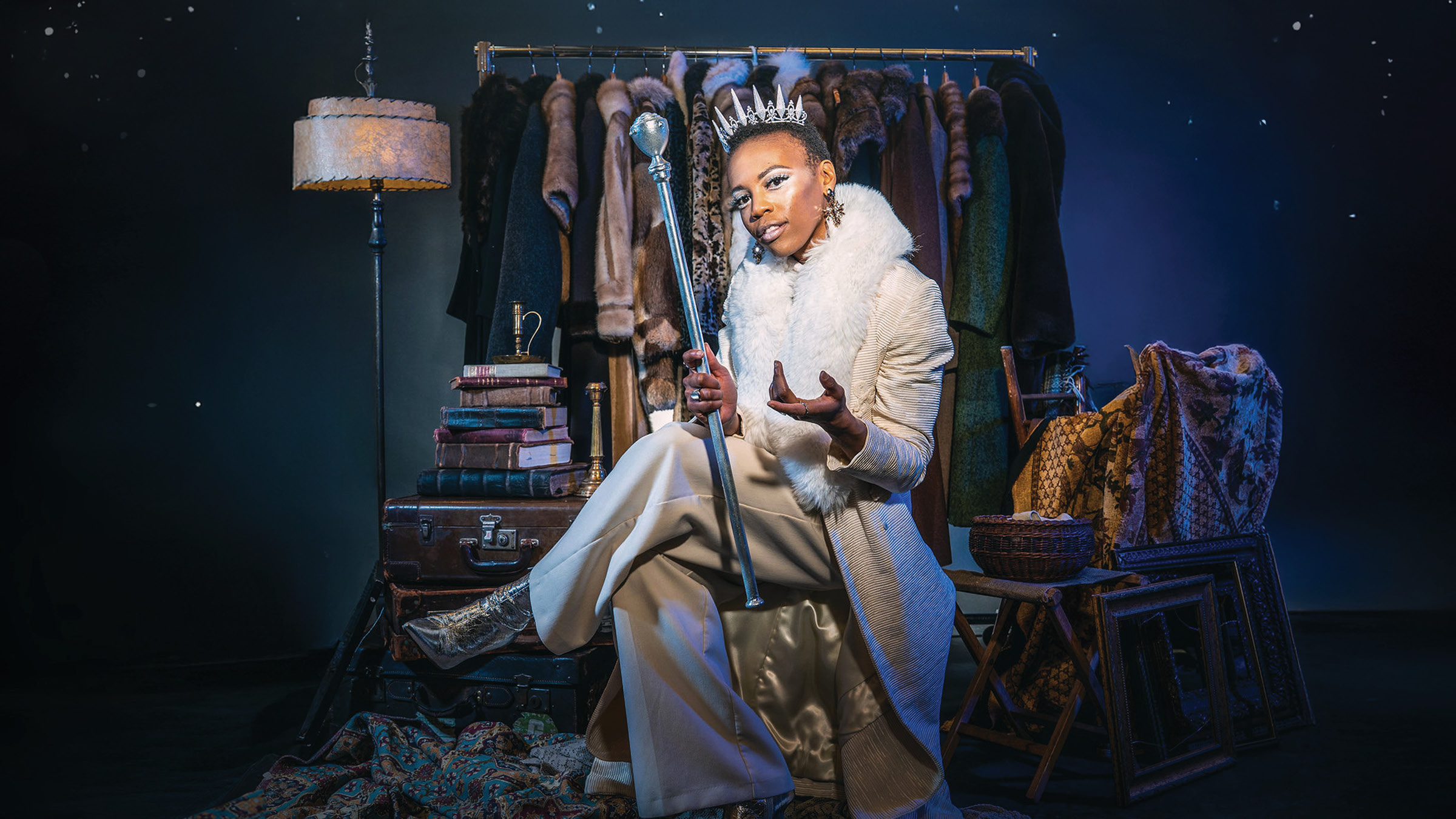 A regal-looking individual dressed in elegant cream and gold attire sits confidently on a vintage suitcase, holding a silver scepter and wearing a jeweled crown. They are surrounded by rich textures: fur coats on a clothing rack behind them, stacked antique books, patterned rugs, and ornate furniture. The lighting casts a dramatic, royal blue hue, emphasizing their powerful, theatrical presence.