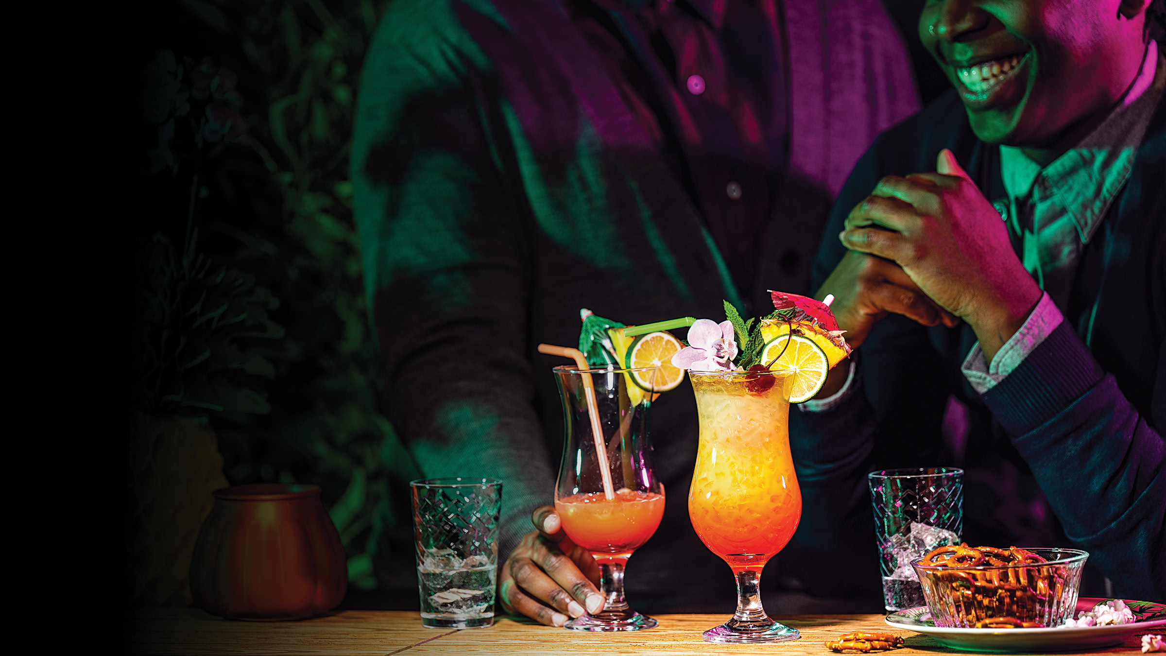 Two people seated at a wooden table in a dimly lit bar, shown from the shoulders down and partially cropped, with coloured green and magenta lighting across their clothing and skin. In the foreground are two tall tropical cocktails in curved glasses, one pink-orange and one yellow-orange, both filled with ice and garnished with lemon slices, a purple orchid, mint, cherries, and small paper umbrellas, with straws in the drinks. A short glass with ice sits on each side of the cocktails, and a bowl of pretzels and a plate with small white snacks are on the right side of the table.