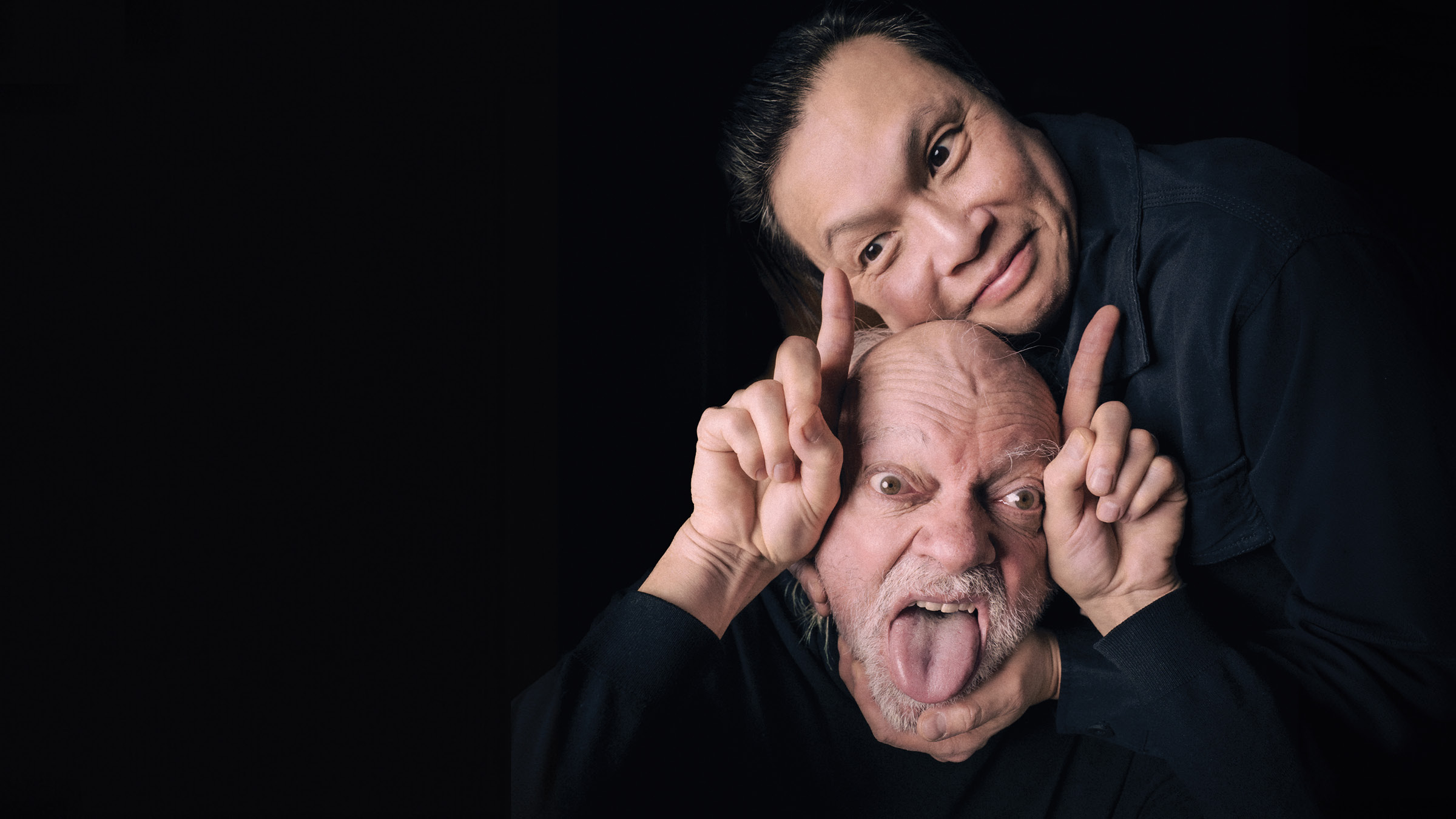 Two people against a black background; the person in front is an older man with a bald head and white beard, making a silly face with his tongue out and eyes wide, while holding up both index fingers like horns. Behind him, a person with long dark hair and a soft smile gently holds the man’s head and rests their own head beside his. Both are dressed in dark clothing.