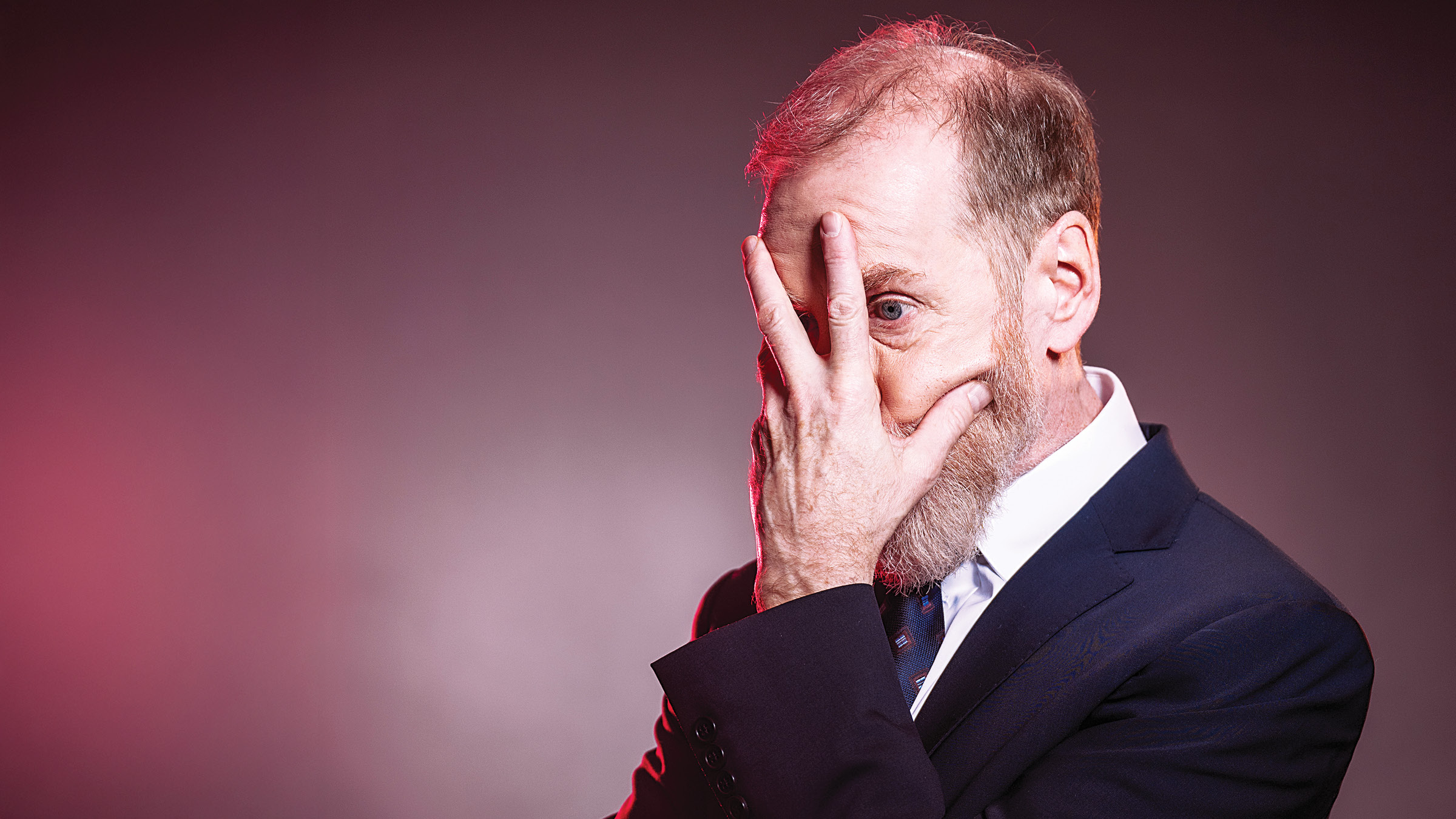 A man in a navy blue suit stands against a gradient background with red and pink lighting. He holds one hand over his face, with fingers spread to reveal one eye looking through. His other arm is crossed under his chest, and he has a short beard and neatly combed hair.