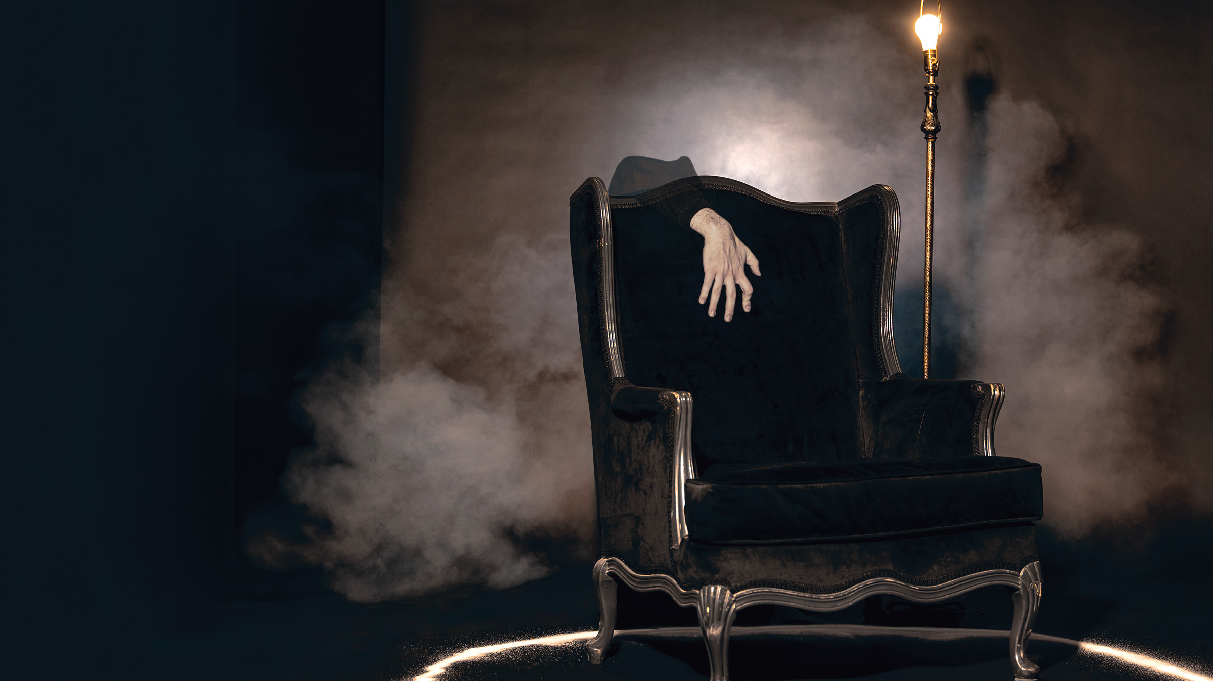 A dark, antique-style armchair with silver trim sits inside a glowing circular outline on the floor, surrounded by mist or smoke. A pale human hand emerges from the back of the chair, appearing to belong to an otherwise invisible figure. A tall, vintage floor lamp with an exposed lightbulb stands to the right of the chair, casting a warm glow in the dim, eerie setting.