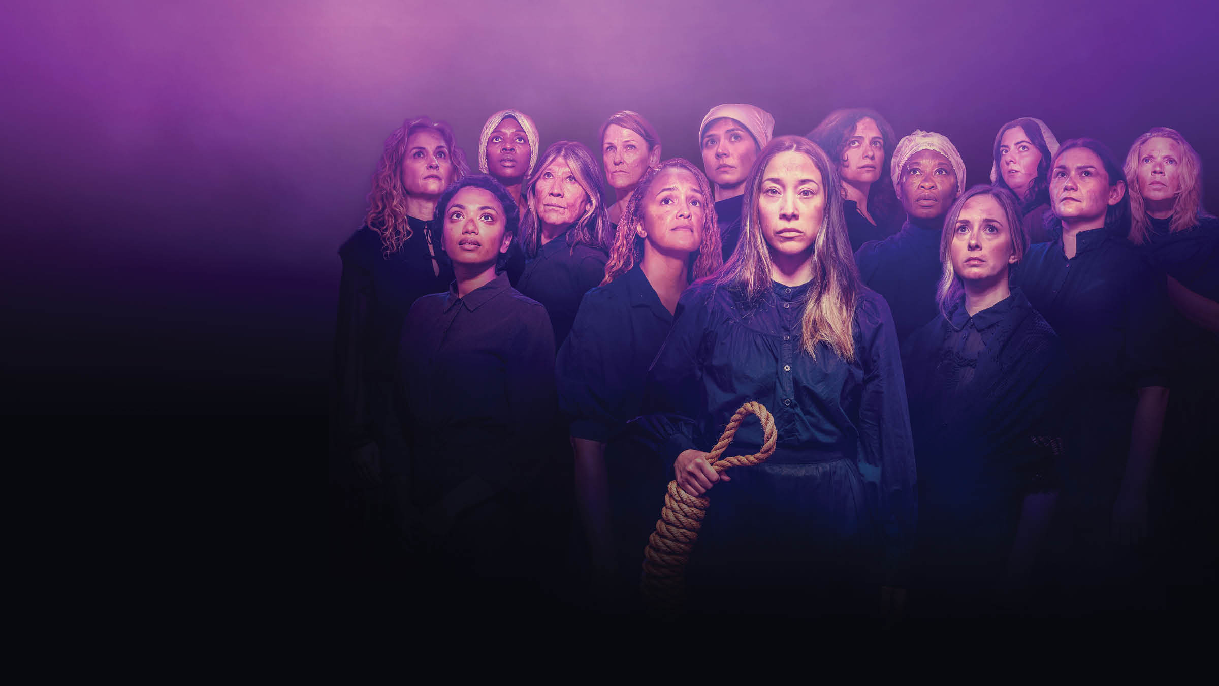 Group of fourteen women of diverse ages and ethnicities dressed in dark, old-fashioned clothing, standing close together and looking upward with serious, solemn expressions. The central woman holds a thick rope in both hands. The image is bathed in dramatic purple lighting with a misty, theatrical atmosphere.