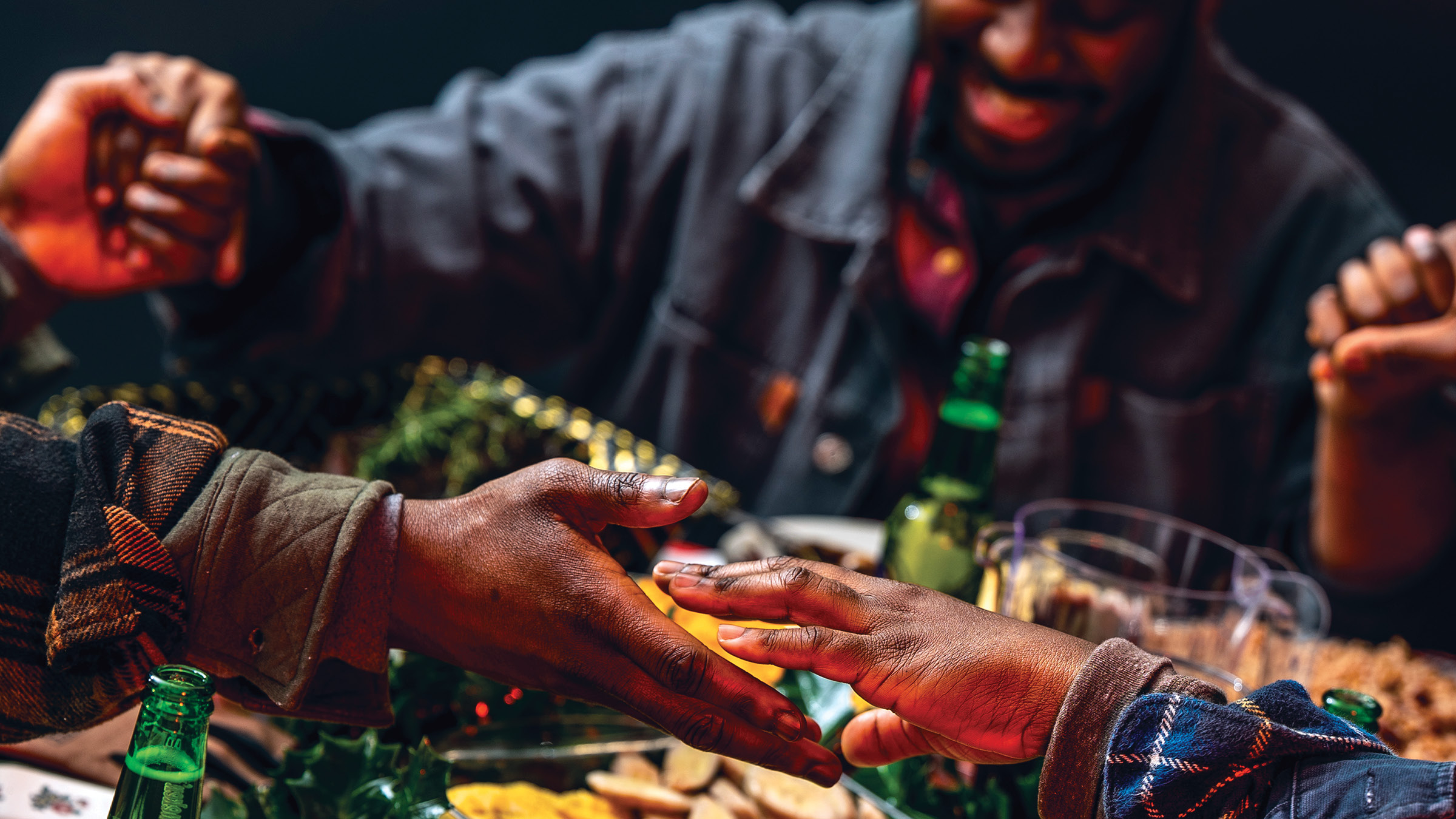 Several people gathered around a table covered with assorted food items such as crackers, pineapple slices, and leafy greens; multiple hands are reaching out in greeting or celebration, and a smiling man in a denim jacket is visible in the background, suggesting a lively and warm social gathering.