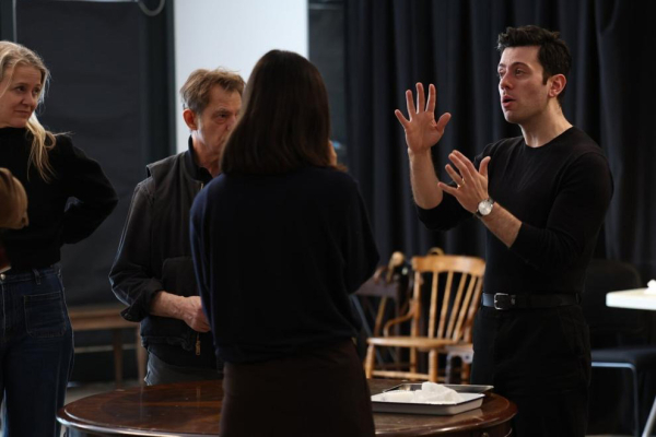 Four people in a room with dark curtains and wooden chairs; one man gestures while speaking, facing a woman seen from behind, as two others observe nearby. A round table with a tray is in the foreground.