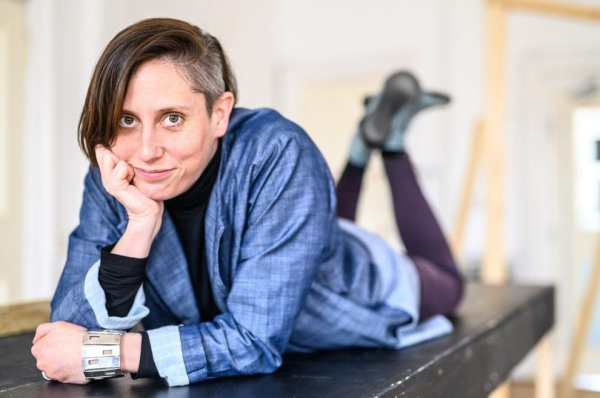 Playwright Erin Shields with short brown hair lying on their stomach on a black platform, head propped on one hand and looking at the camera with a slight smile. They are wearing a blue jacket over a black top, purple leggings, and dark shoes, with a wide metallic bracelet on one wrist; the indoor background is softly blurred.