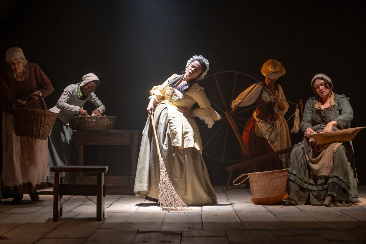 Five women in historical clothing are engaged in domestic tasks under stage lighting. One woman in the centre wears a yellow dress and holds a broom while leaning to one side in apparent discomfort. Others wash, spin, or sift items, with a large spinning wheel and baskets nearby.