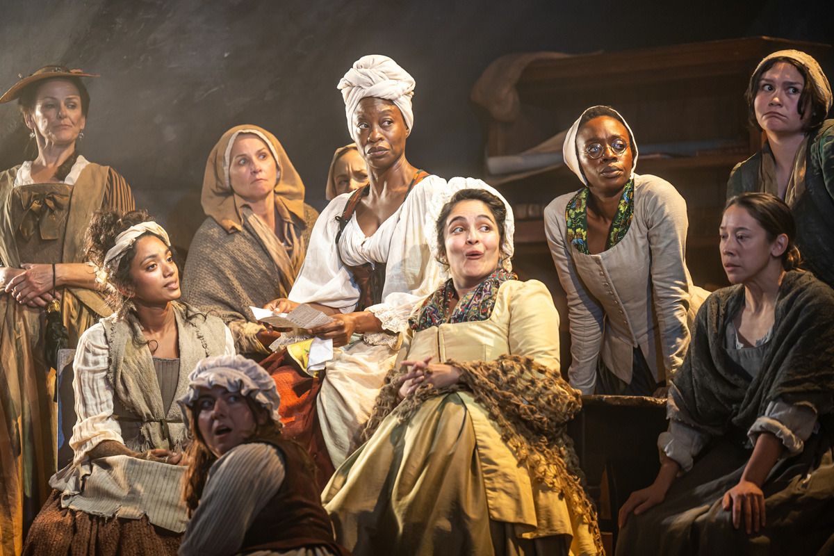 A group of women in 18th-century-style clothing are gathered closely together indoors under warm stage lighting. The central woman in a light yellow dress and patterned shawl has an animated expression, while the others around her exhibit a mix of attentiveness, concern, and curiosity. One woman in white holds a piece of paper, and a wooden cabinet with folded linens is visible in the background.