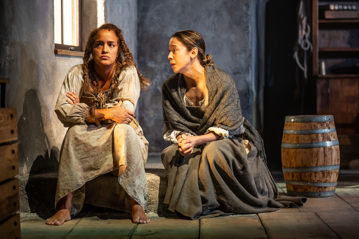 Two women in period clothing sit on a low stone ledge in a dimly lit, rustic interior. The woman on the left, barefoot and wearing a worn light-coloured dress with shackles on one wrist, stares forward with a serious expression and crossed arms. The woman on the right, wrapped in a grey shawl, leans in as if speaking gently. A wooden barrel and dark wooden shelves are visible in the background.