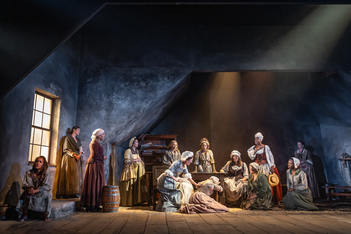 Thirteen women in 18th-century clothing are positioned across a dimly lit wooden stage with a sloped ceiling. At the centre, one woman kneels beside another who is collapsed on the floor, clutching her torso. Others stand or sit around a large wooden table, observing the scene with concern or focus. A few women stand near a window at the left, illuminated by soft light, while another group gathers near a cabinet and a spinning wheel on the right. A beam of light falls dramatically on the central figures, emphasizing the tense moment.
