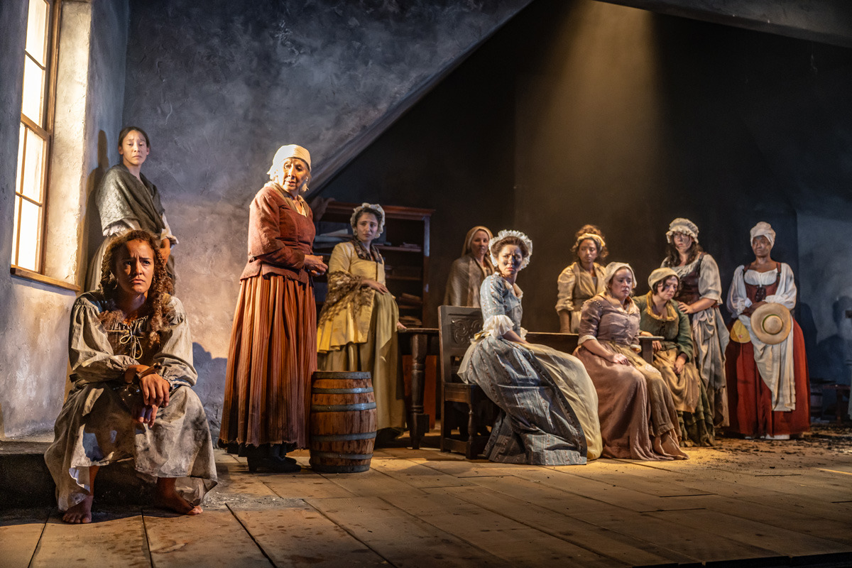 A group of eleven women in 18th-century clothing are gathered on a dimly lit wooden stage. One woman sits barefoot near a window at the far left, with a solemn expression and shackles on her wrists. The others are positioned across the room—some standing, some seated—most looking toward the centre with serious or contemplative expressions. The lighting casts a soft glow from the window and a focused beam from above, enhancing the tense, quiet atmosphere.