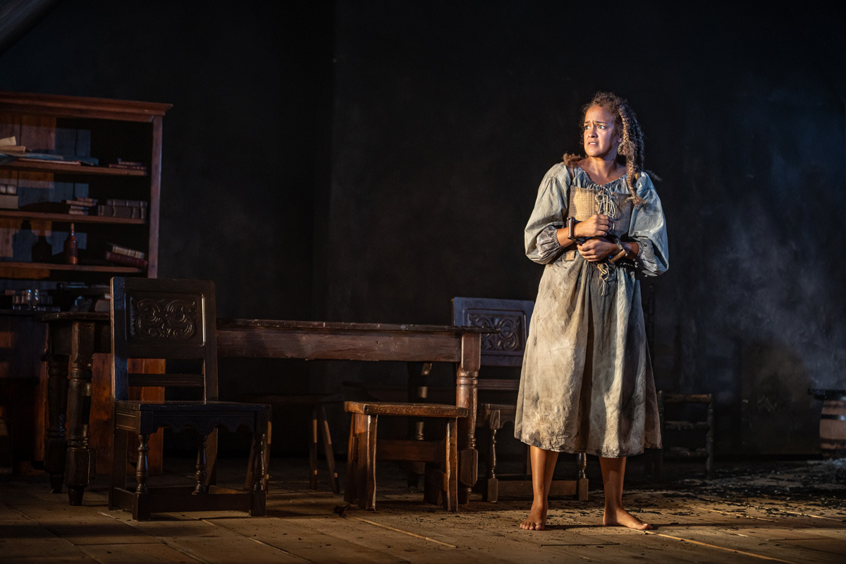 A barefoot woman in a worn, laced dress stands alone on a wooden stage, holding a small bundle to her chest. Her wrists are in shackles, and she gazes off to the side with a distressed expression. The dimly lit room behind her contains a large wooden table, carved chairs, and a bookshelf filled with scattered books and bottles. The lighting casts deep shadows, emphasizing her isolation.