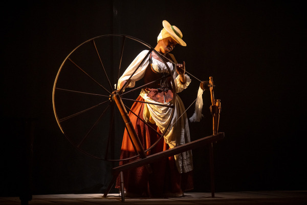 A woman in a wide-brimmed hat and 18th-century attire operates a large spinning wheel on a dimly lit stage, her hand pulling thread as warm light highlights her figure against a dark background.