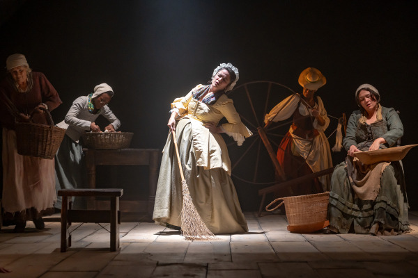 Five women in historical clothing are engaged in domestic tasks under stage lighting. One woman in the centre wears a yellow dress and holds a broom while leaning to one side in apparent discomfort. Others wash, spin, or sift items, with a large spinning wheel and baskets nearby.