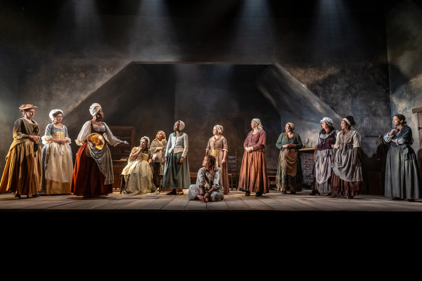 Thirteen women in 18th-century clothing are gathered across a wide wooden stage beneath dramatic overhead lighting. Thirteen stand or sit in a semi-circle, facing a barefoot woman seated cross-legged on the floor at the centre, her hands in shackles. Their expressions range from serious to contemplative, and the setting features dark textured walls, a slanted ceiling, and sparse period furnishings including a brick hearth and wooden shelves.
