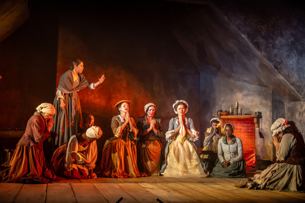 A group of women in 18th-century clothing are gathered in a dimly lit interior with warm, reddish lighting, suggesting firelight. Most of the women are kneeling or sitting on the wooden floor in prayer, hands clasped and heads bowed. One woman stands to the left with an arm raised, appearing to address the group. A red-bricked hearth with candles is visible in the background, contributing to the dramatic, solemn atmosphere.