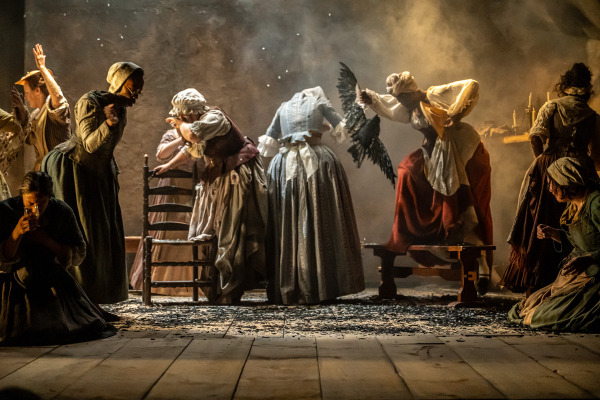 A group of women in period clothing are positioned across a wooden stage, with dramatic lighting and theatrical smoke. Most are dressed in long skirts, aprons, and bonnets, evoking an 18th or 19th-century setting. One woman in the centre raises a black bird, possibly a raven, while others react in various expressive poses—some appear distressed, others shocked or contemplative. A few women kneel or crouch near the front, and a table with lit candles is visible in the background to the right. The atmosphere is tense and dramatic.