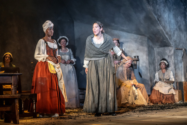 A woman in a grey shawl and long dress stands at the centre of a dimly lit stage, extending one arm as she speaks with urgency. To her left, another woman in a white blouse and red skirt stands silently, watching with a serious expression. Several women in period clothing sit or stand in the background against dark, textured walls, some appearing concerned or exhausted. The wooden floor is scattered with black debris.