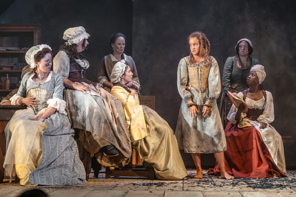 A barefoot woman in a faded dress stands at the centre of a wooden stage with her hands in shackles, held low in front of her. Her expression is tense as several women in 18th-century clothing sit or stand around her, watching intently with mixed expressions of concern, curiosity, or disapproval. The stage floor is scattered with dark debris, and a wooden shelf with bottles is visible in the background.