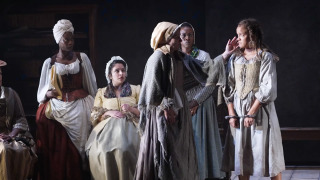Six women dressed in historical clothing are on stage in a dramatic scene. One woman in the centre, wearing a bonnet and grey shawl, reaches out to touch the face of a younger woman in shackles, who looks distressed. The others watch intently, with expressions ranging from concern to curiosity. The lighting is dim, creating a serious atmosphere, and the background is dark with minimal set pieces.