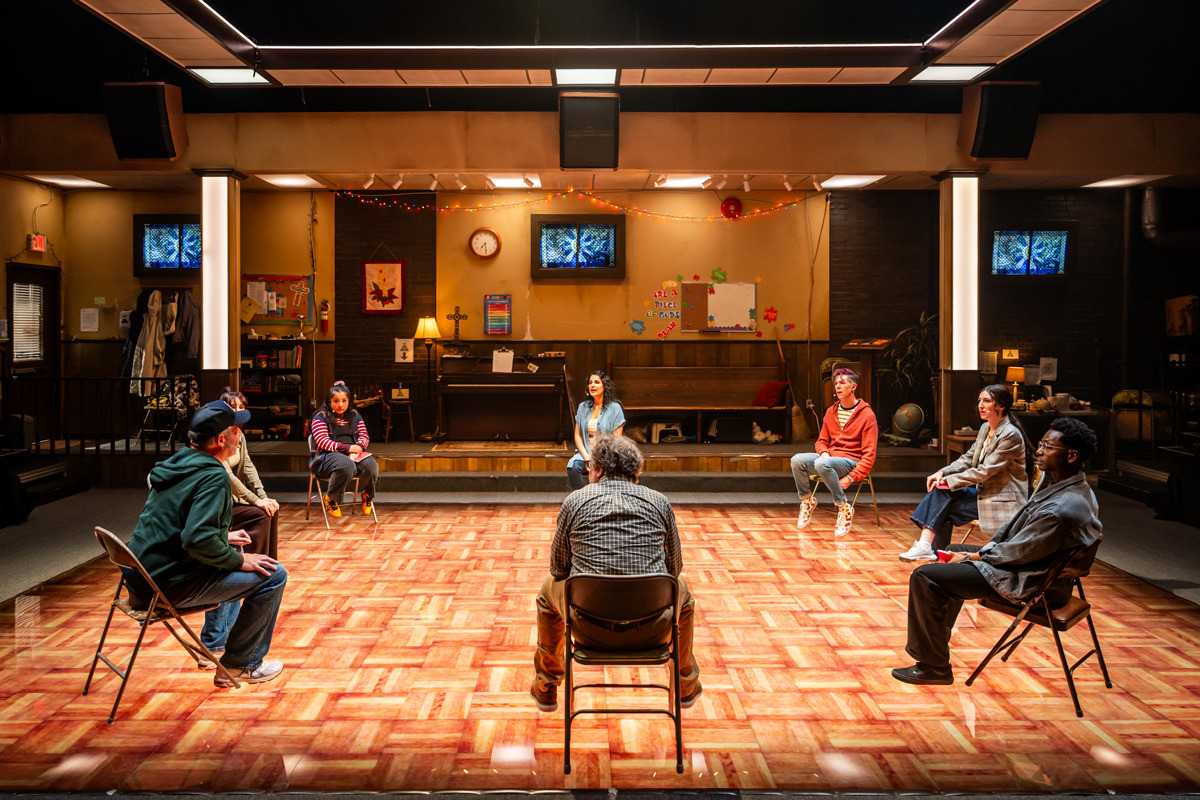 A group of eight people sit in a circle on folding chairs in a brightly lit room with a wood-patterned floor, resembling a community or church hall. The background features a stage area with a piano, a bulletin board, a wall clock, decorative string lights, and various posters and crafts on the walls. The room includes elements like a coat rack, bookshelves, and a globe, suggesting a multipurpose space. The individuals appear engaged, possibly participating in a discussion or support group.