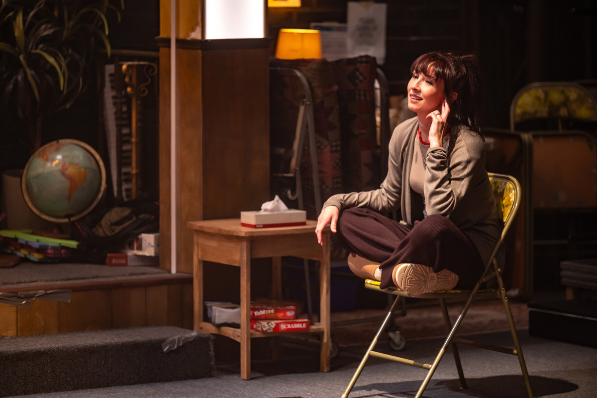 A woman sits cross-legged on a folding chair, smiling with one hand resting near her ear as if listening. She wears relaxed, casual clothing and is bathed in warm lighting. Beside her is a small wooden table with a tissue box on top and board games stored underneath, including Scrabble and Yahtzee. In the background of the church basement setting are stacked chairs, a globe, and various supplies.