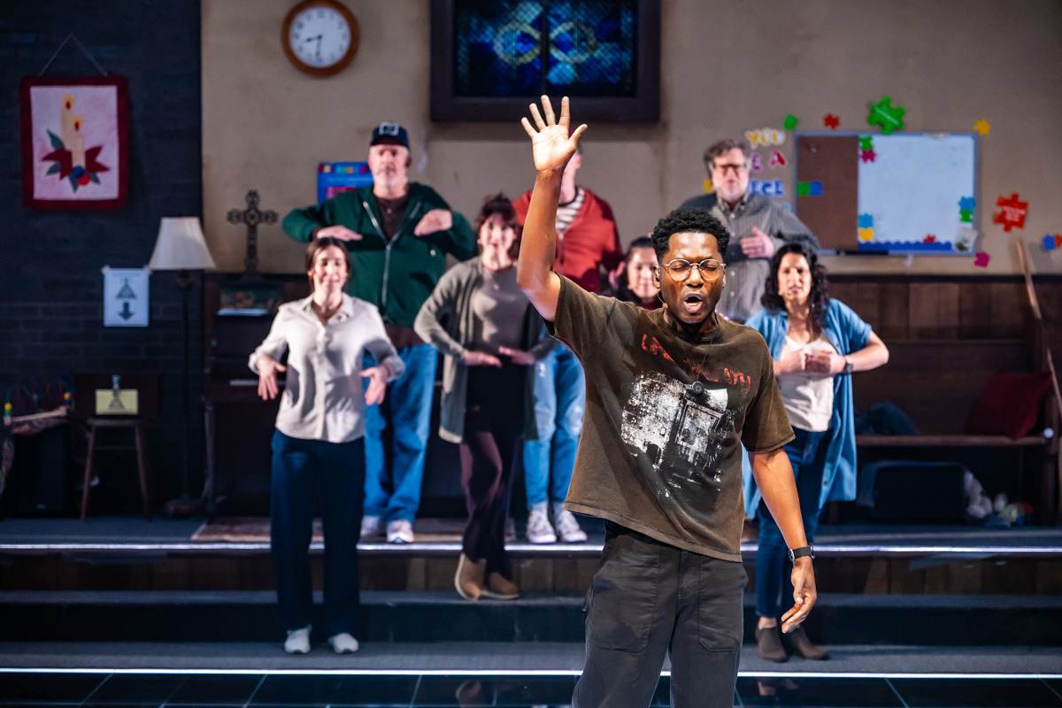 A man stands at the front singing with one arm raised and an expressive face. Behind him, seven other people stand in a staggered formation with their hands positioned in front of their chests, facing forward. The setting is a church basement with a piano, bulletin board, clock, and colourful decorations visible on the back wall.