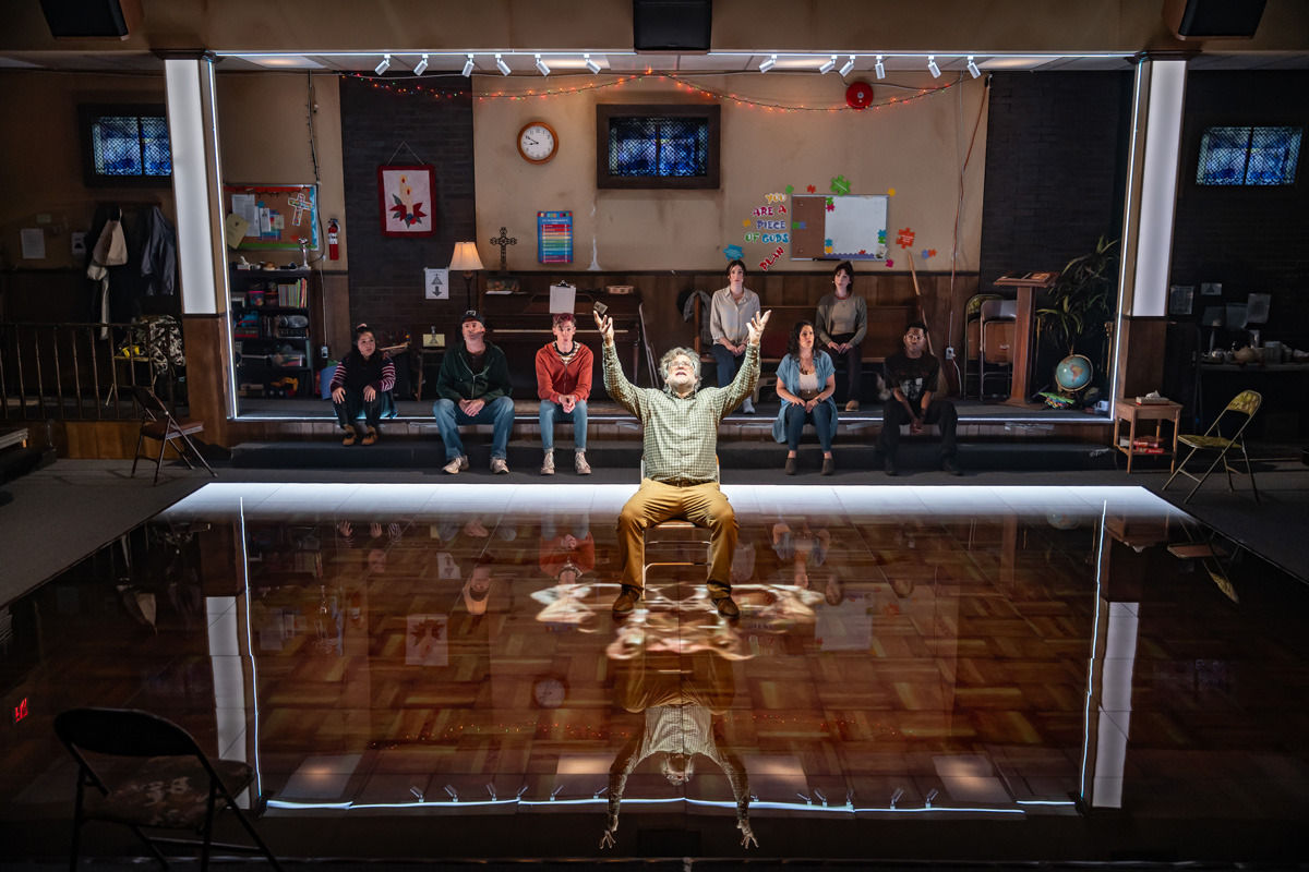 A man sits alone at the centre of a highly reflective floor with his arms raised and face lifted, framed by a glowing square of light. Behind him, eight people sit in a row on risers against the wall of a church basement. The background includes a piano, bulletin boards, religious decorations, a globe, and various signs and crafts, creating a warm, lived-in environment. The man’s reflection is clearly visible on the glossy floor beneath him.