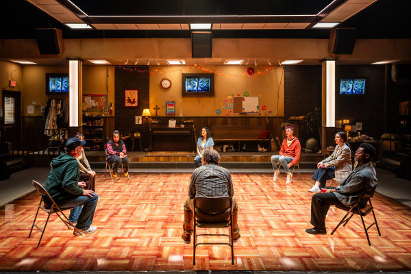 A group of eight people sit in a circle on folding chairs in a brightly lit room with a wood-patterned floor, resembling a community or church hall. The background features a stage area with a piano, a bulletin board, a wall clock, decorative string lights, and various posters and crafts on the walls. The room includes elements like a coat rack, bookshelves, and a globe, suggesting a multipurpose space. The individuals appear engaged, possibly participating in a discussion or support group.