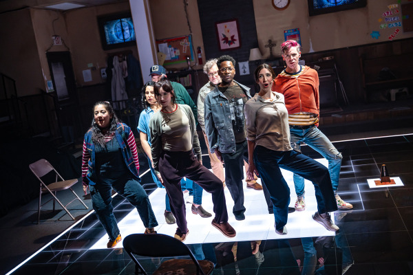 Eight people stand in a diagonal formation on a glowing white square in the centre of a dark, reflective floor. Each person has one leg raised in a stylized pose, and they appear to be mid-performance with expressive facial expressions. The group wears casual clothing, and the background reveals a church basement with folding chairs, a piano, bulletin boards, and religious-themed decorations. A metronome sits on the floor to the right.